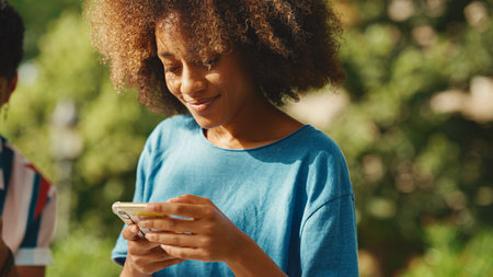 Close-up of young smiling woman with curly hair sitting in park having picnic with friends on summer day outdoors. Girl uses cellphone, browse social networks, photo, videoの写真素材