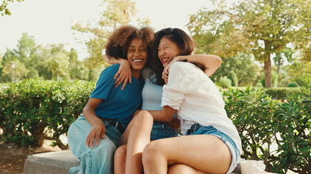 Happy multiethnic young women talking while sitting on park bench on summer day outdoors. Group of girls talking and laughing merrily in city parkの写真素材