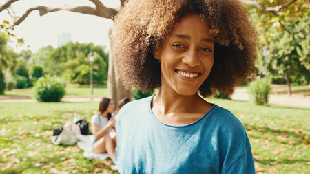 Portrait of young smiling woman with curly hair wearing blue t-shirt posing for the camera in the park . Picnic on summer day outdoors her friends sitting in distance blurred on backgroundの写真素材