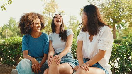 Happy multiethnic young women talking while sitting on park bench on summer day outdoors. Group of girls talking and laughing merrily in city parkの写真素材