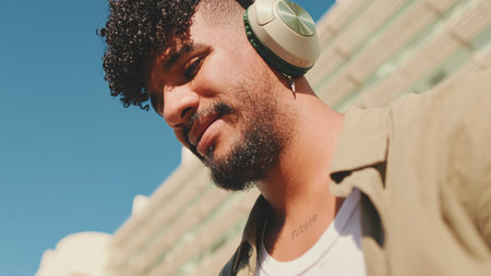 Close up, young male student dressed in an olive color shirt sits outside next to the university, listens to music on headphones, selects tracks on his phoneの写真素材