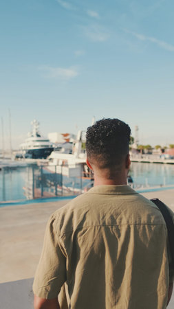 Young man dressed in an olive-colored shirt is standing in the port looking at the yachts. back view