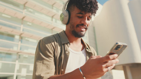 Close up, young male student dressed in an olive color shirt sits outside next to the university, listens to music on headphones, selects tracks on his phoneの写真素材