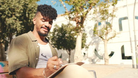 Young male student wearing an olive colored shirt takes notes in notepad while sitting on bench outside the universityの写真素材