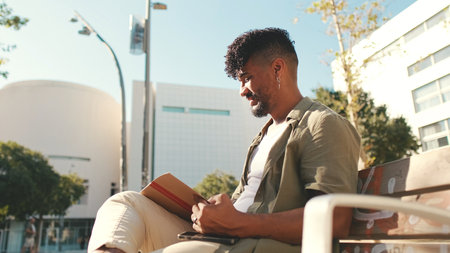 Young male student wearing an olive colored shirt takes notes in notepad while sitting on bench outside the universityの写真素材