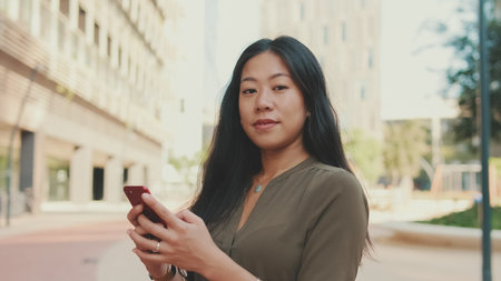 Close-up of young happy businesswoman using mobile phone while standing on modern buildings backgroundの写真素材