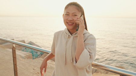 Smiling Asian girl in sportswear talking on the phone while standing on the embankment on modern background buildings in the morning lightの写真素材