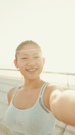 Smiling asian girl in sports top makes selfie while standing on the embankment in the morning lightの写真素材