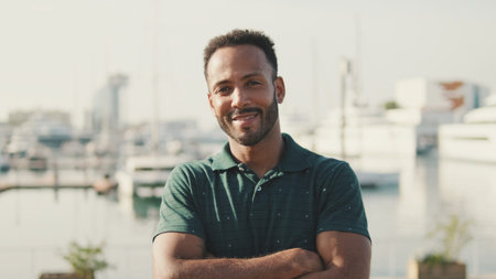 Young man sitting in the harbor with yachts on the background, looking at the camera and smiling