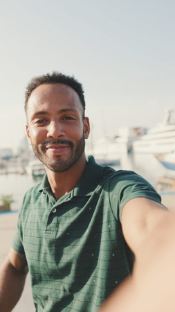 Happy young man making video call from phone while sitting in harbor with yachts on background