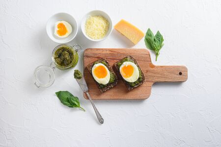 Fresh Green basil pesto in glass jar homemade on old rustic wood eggs panini bread with pesto knife and hot sriracha sauce italian breakfast with ingredients green pesto top view on white concrete table.の写真素材