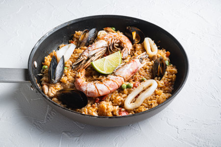 Seafood and chicken paella with rice, mussles, shrimps, chicken, tomatoes and wine in pan on white textured background, food photo.の写真素材