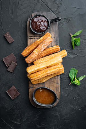 Typical Spanish snack churros, fried-dough pastry served usually with chocolate caramel hot sauce set, on black background, top view flat layの写真素材