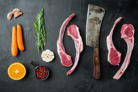 Frenched raw fatty lamb chops set, with ingredients carrot orange, herbs, on black stone background,...