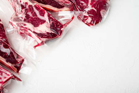 Fresh tasty raw beef steaks with bone wrapped in vacuum plastic packing at market set, tomahawk, t bone, club steak, rib eye and tenderloin cuts, on white stone background, with copy space for textの写真素材