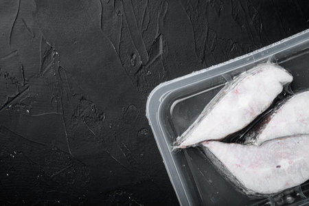Frozen Greenland halibut steaks vacuum pack set, on black dark stone table background, top view flat...