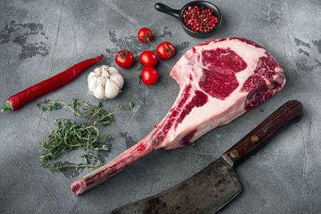 Dry aged raw tomahawk beef steak with ingredients for grilling set, and old butcher cleaver knife, with seasoning and herbs, on gray stone backgroundの写真素材