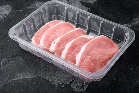 Raw organic meat. Pork steaks, fillets for grilling, baking or frying set, in plastic pack container, on black dark stone table background, with copy space for textの写真素材