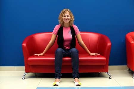 Portrait of young woman sitting on red sofa against blue wallの写真素材