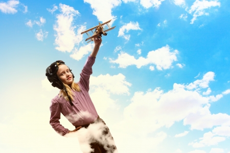Image of little girl in pilots helmet playing with toy airplane against clouds backgroundの写真素材