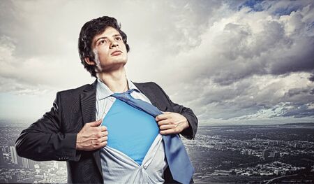 Image of young businessman showing superhero suit underneath his shirt standing against city backgroundの写真素材