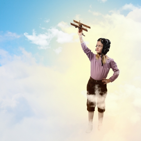 Image of little girl in pilots helmet playing with toy airplane against clouds backgroundの写真素材