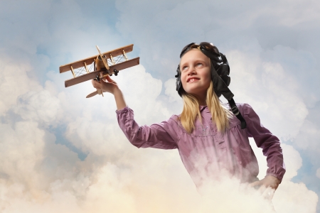Image of little girl in pilots helmet playing with toy airplane against clouds backgroundの写真素材