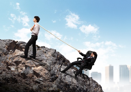 Image of two young businessmen pulling rope atop of mountain against city backgroundの写真素材