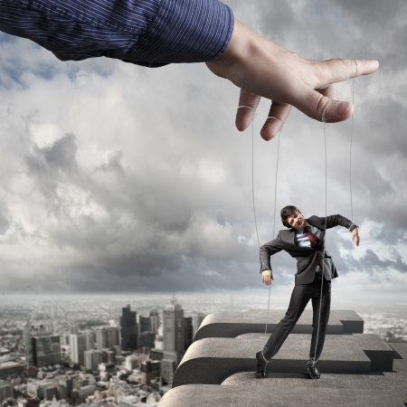 Businessman marionette on ropes controlled by puppeteer against city backgroundの写真素材