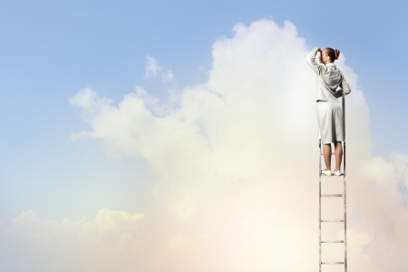 Businesswoman standing on ladder looking into distance against cloudy backgroundの写真素材