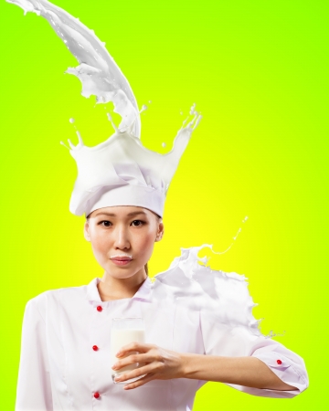 Asian female cook against milk splashes in red apron against color background holding glass of milkの写真素材