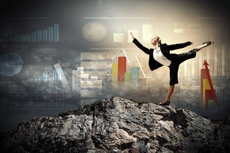 young businesswoman standing on one leg atop of mountain against diagram backgroundの写真素材