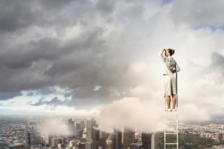 Businesswoman standing on ladder looking into distance against city backgroundの写真素材