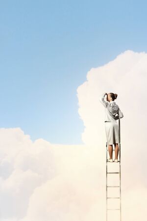 Businesswoman standing on ladder looking into distance against cloudy backgroundの写真素材