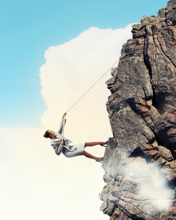 businesswoman climbing steep mountain hanging on ropeの写真素材