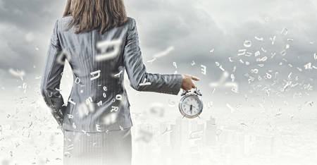Back view of businesswoman holding alarm clock against city backgroundの写真素材