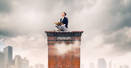 Young attractive businesswoman sitting on roof of building and reading bookの写真素材