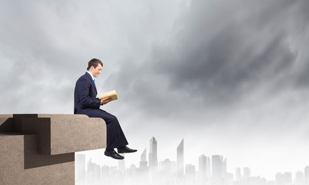Young businessman sitting on top of building and reading bookの写真素材