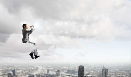 Young carefree businessman sitting on cloud and playing fluteの写真素材