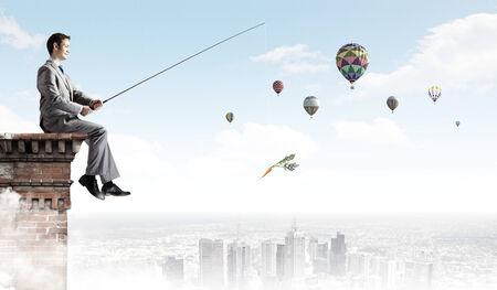 Businessman sitting on top of building and fishing with rodの写真素材