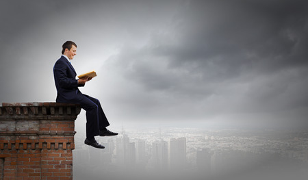 Young businessman sitting on top of building and reading bookの写真素材