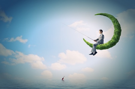 Businessman sitting on green moon and fishing with rodの写真素材