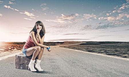 Young pretty girl traveler sitting on suitcase aside of roadの写真素材