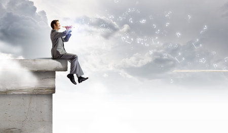 Young carefree businessman sitting on top of building and playing fluteの写真素材
