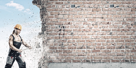 Strong man in uniform breaking brick wall with hammerの写真素材