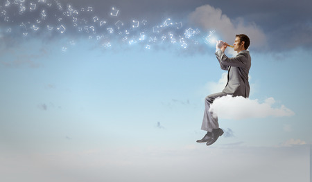 Young carefree businessman sitting on cloud and playing fluteの写真素材