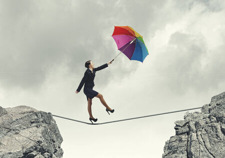 Young businesswoman walking on rope above gap with colorful umbrellaの写真素材