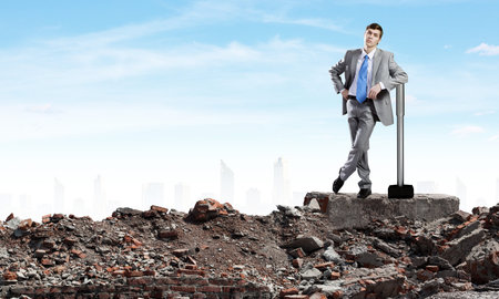 Young businessman with hammer standing on ruinsの写真素材
