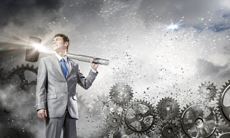 Young determined businessman with hammer on shoulder and cogwheels at backgroundの写真素材