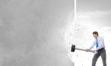 Young determined businessman with big hammer in hands crashing wall